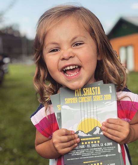 Young girl laughing with a handful of Mt. Shasta Community Concert Series postcards in her hand.